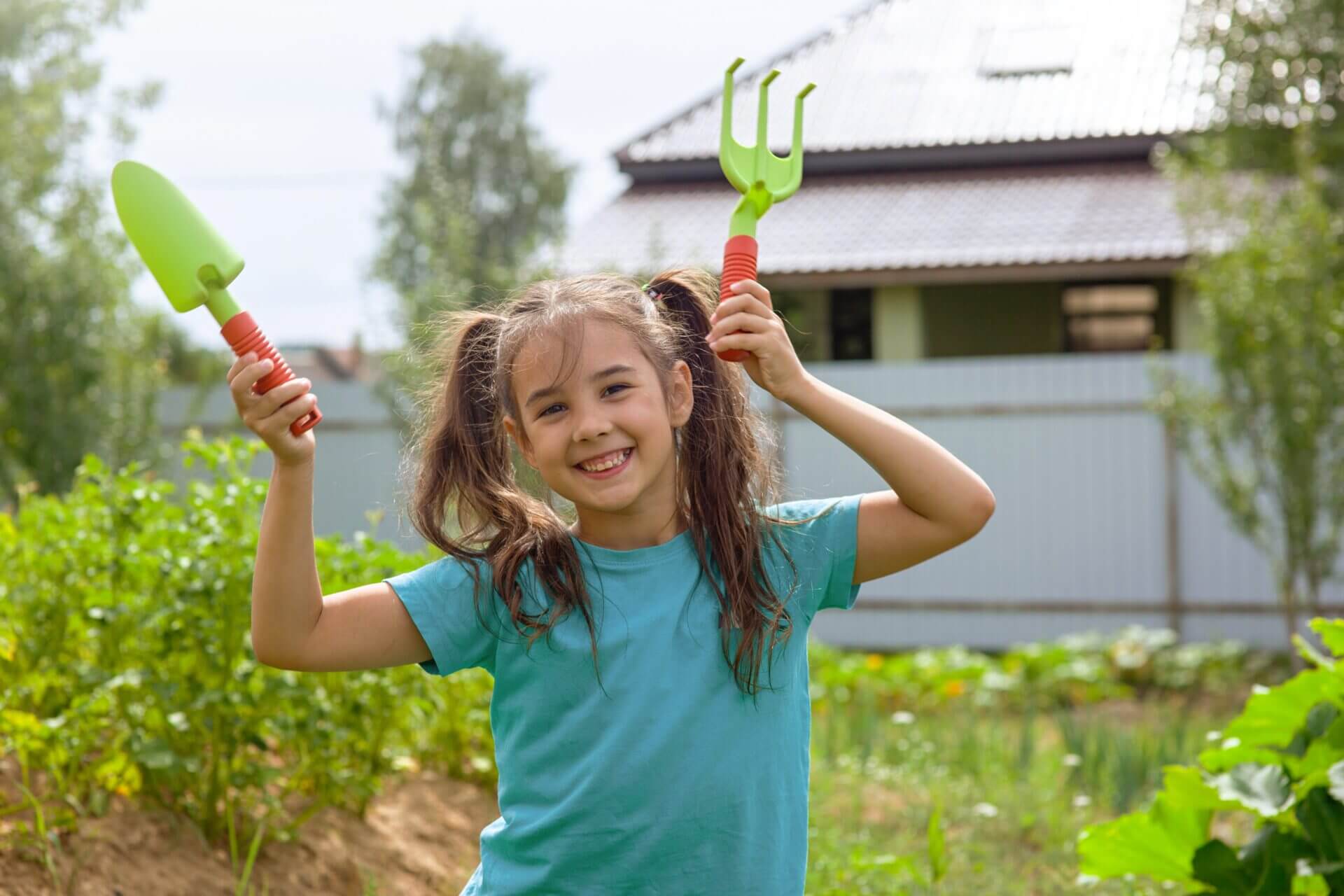 A happy girl holding gardening tools in a backyard.
